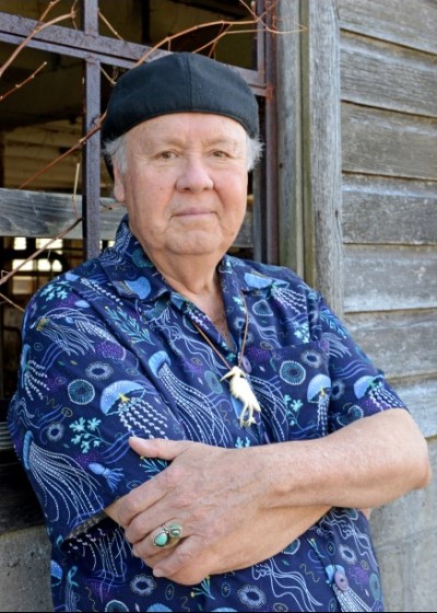 Person with crossed arms leaning against a window of a wooden structure wearing a blue shirt with a jellyfish design and a black cap over white hair.