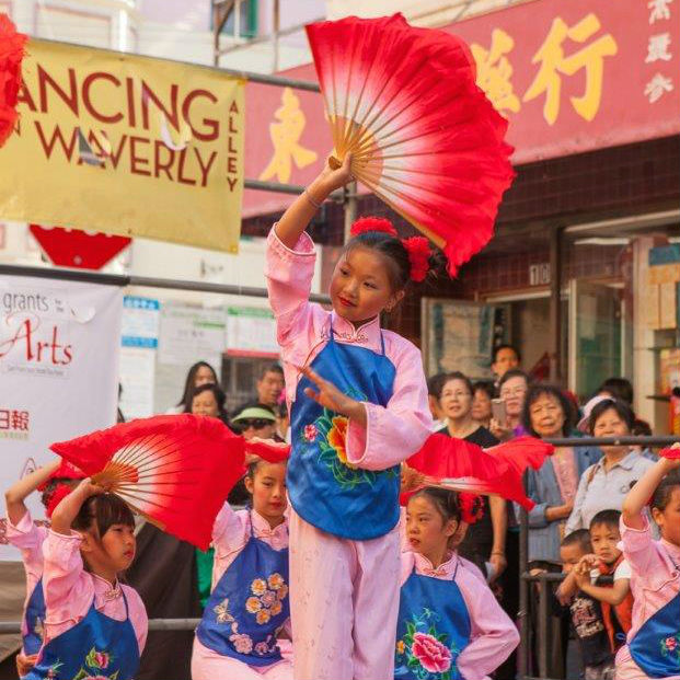  Young children with matching pink costumes, blue floral aprons, and red fans, dance on an outdoor stage.
