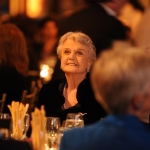 Smiling person sitting among people (blurred) wearing a black dress and white pearls. 