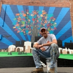 Greg Puckett wears paint-stained clothes and sits in front of a bright painted mural depicting a tree where the leaves are handprints.