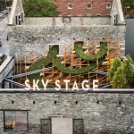 A photo from above of a gray stone building with an open rooftop area featuring trees and other plants and the words “Sky Stage” on the roof edge.