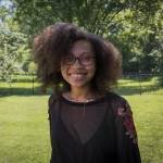 A smiling young Black woman wearing glasses, dark curly hair, and a black top with embroidered floral sleeves.