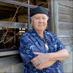 Person leaning against a building with wooden siding, arms crossed, wearing a black hat, blue pattered short-sleeved shirt, necklace with a white bird pendant, and a turquoise ring. 