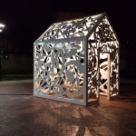 A nighttime photo of a public art sculpture in the shape of a small house covered in decorative cut-out shapes. A light from inside casts shadows of the shapes around the sculpture.