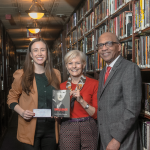 Regina Burgher, Jeanne-Marie Osterman, and Nolen Bivens standing smiling in a long hall full of books. Burgher is holding a check, and Osterman is holding her book, Shellback.