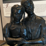 A bronze sculpture of two figures sitting side by side leaning on each other and holding hands.