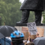 It's a photo of a protest at the foot of a large public art monument. In the foreground a protestor holds up a sign that reads "Racism is a pandemic too." 