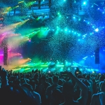It's a photo of a large crowd at a concert, with rainbow colors lighting up the stage.