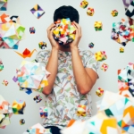 Person in a light colored shirt against a white background holding and surrounded by colorful 3 dimensional shape objects.