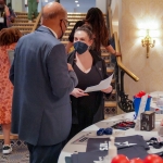 At a conference, a person sits at a table piled with swag listening to two standing people having a conversation. All three wear business casual attire and masks.