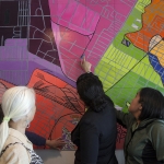People look and point at a large colorful mural painted to resemble a neighborhood street map.
