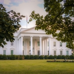 Photo of the White House, a large white neoclassical building, with leafy green trees framing the view