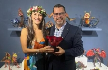 Two people stand smiling in front of an art display. One wears a lei around their head and is accepting an award from the other.