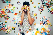 Person in a light colored shirt against a white background holding and surrounded by colorful 3 dimensional shape objects.