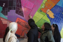 People look and point at a large colorful mural painted to resemble a neighborhood street map.