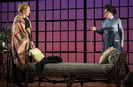 Two actors on stage before a purple backdrop. One is wearing a blue dress and holding a gold chain and the other is wrapped in a blanket. There is a bench with pillows in front of them. 