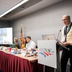 Three people sit at a table and one stands at a podium as panelists or hosts for a meeting.